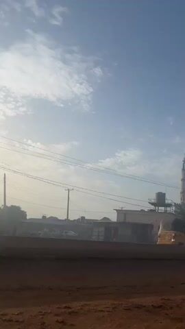 The video shows a street scene with a mosque in the background. The mosque has two minarets and a green dome. There are buildings and vehicles on the street, including a yellow auto rickshaw and a red car. People can be seen walking on the sidewalk. The road is unpaved and has some puddles. The sky is blue with some clouds. There are power lines overhead.