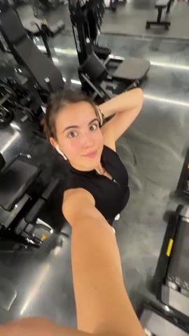 A young woman with brown hair tied back is taking a selfie video in a gym. She is wearing a black sleeveless top and black leggings. She has white earbuds in and is smiling at the camera. The gym appears to have treadmills and other exercise equipment. The video is a bit blurry due to motion. She waves at the camera in the second clip and smiles in the third clip.