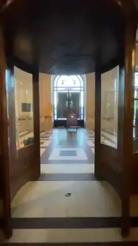 The video shows a view of an interior space, likely a hallway or lobby. The camera moves slowly from a doorway into the space. The floor is covered with a pattern of alternating white and brown squares. There are large arched windows on the left side of the hallway, revealing people standing outside. The walls are beige and adorned with plaques and other decorations.  There are ornate hanging lamps providing light. People can be seen walking and standing in the hallway, interacting with each other. The space appears to be part of a grand or historic building.