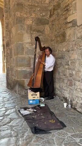The video shows a street performer in Florence, Italy, playing the harp in a stone archway. The man stands to the left side of the screen wearing a white button up shirt and black pants. He stands in front of a large wooden harp, which rests upon a cardboard box. In front of him on the ground is a blanket with money on it, including coins and bills. To the left of the blanket is a disposable cup and a water bottle. The archway is made of weathered stone, with an opening to the street visible on the left side of the screen.
