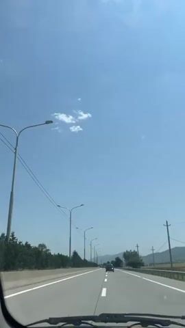 The video shows a view from inside a vehicle traveling on a highway. The sky is bright blue with a few scattered clouds. On the left side of the highway, there is a line of trees and a median barrier. Streetlights are spaced along the side of the road. On the right side, there are more trees and some utility poles. Several cars are visible on the highway ahead, all moving in the same direction as the camera. The road is paved and has white lane markings. The overall impression is of a clear, sunny day on a highway.