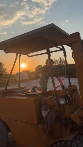 A young boy is exploring the driver's area of a yellow forklift. He is kneeling on the seat and reaching for the steering wheel. The forklift is silhouetted against a sunset, with the sun visible through the forklift's protective cage. A small white van is parked in the background.