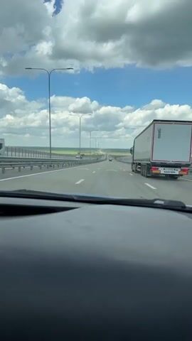 The video shows a view from the driver's seat of a car traveling on a highway. The sky is blue with scattered white clouds.  To the right of the car is a large white semi-truck.  The road is multi-laned and divided by a center barrier.  There are lamp posts lining the side of the highway. Distant cars can be seen ahead on the road. A green field stretches out on either side of the highway.  The dashboard of the car is visible at the bottom of the frame.