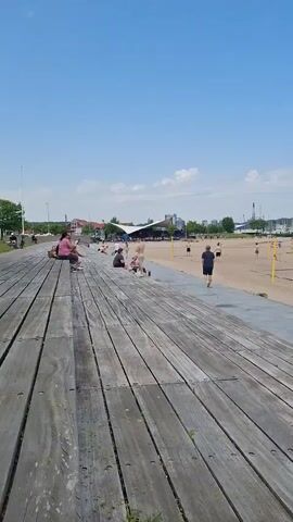The video shows a sunny day at a beach with a wooden boardwalk in the foreground. People are scattered along the beach, some sunbathing and others playing volleyball. In the background, there is a body of water with boats visible. A few people are sitting on the boardwalk.