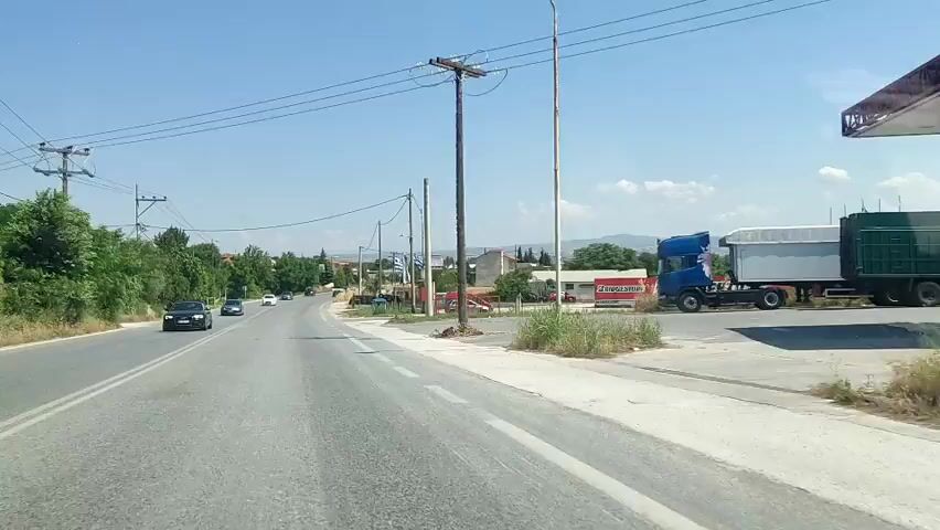 The video captures a forward-facing view from a vehicle driving on a paved two-lane road under a clear blue sky. On the left, several cars are visible driving in the opposite direction, and green trees line the roadside. Utility poles with power lines run alongside the road. On the right, a Bridgestone sign is visible initially. As the vehicle moves forward, two Greek flags on poles come into view. Further along, a green trash bin is seen on the right, followed by a property with a red fence and stacks of tires. Hills are visible in the distance.