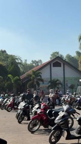The video shows a parking lot filled with parked motorcycles under a clear blue sky. In the background, there is a building with a brown roof and a white facade adorned with colorful murals. Several people are standing and interacting in front of the building, some are wearing headscarves. Palm trees and other greenery surround the area, suggesting a warm, possibly tropical, climate. The motorcycles are parked in rows, and some are covered with helmets. The overall impression is a busy outdoor scene with people arriving or departing from the building.
