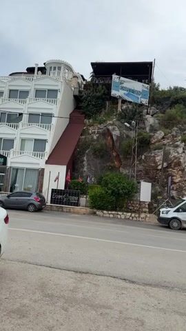 The video captures a roadside scene in Finike, Turkey, under an overcast sky. It begins by showing a multi-story white building with multiple windows and a distinctive, inclined, reddish-brown ramp or walkway extending upwards along its side. A sign near the base of this building reads "CARPE DIEM GOLD". Two cars are parked close to the building. Higher up on the rocky hillside behind, another sign for "CARPE DIEM" is visible.

As the camera pans to the right, a white commercial van is seen parked on the shoulder of the road. Its side is marked with "DIAMOND PERFORMANCE OKAYTAS". The background is dominated by a large, rugged rocky hillside, characterized by distinct layers of rock and sparse green shrubs and trees. The sky is uniformly grey and cloudy.

Continuing the pan, a white sedan with "i.kilican" written on its rear window comes into view, parked on the side of the road. Behind it, a light green sedan is also parked. Next to these cars, a small, white, simple structure is visible, featuring a satellite dish and a sign that reads "FERDİN'İN YERİ". A partially visible red sign with the word "KÖFTE" is also seen in this area. The road occupies the foreground throughout the video. No people are visible.