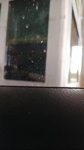 From inside a car, the camera focuses on the rain-spattered glass of a building. A person wearing an orange and yellow safety vest walks behind the glass. The person is blurry and out of focus.  The interior of the car includes a textured black surface near the bottom of the frame. The exterior of the building is white.