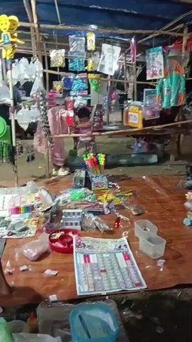 This is a video of a market stall. In the background, there is a woman in a pink dress. In front of her are various toys and other items displayed on shelves. In the foreground, there are more toys, including bubble blowers, small cars, and other trinkets, spread out on the ground. There is also a calendar or chart with what appear to be small images. Two children are present in the scene. The stall is covered by a tarp, and the ground is covered with a mat. The overall impression is a chaotic but lively market setting.