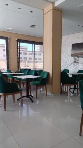 The video pans across a cafe with multiple small tables and green upholstered chairs. Large windows overlook buildings and an outdoor area. A laptop is visible in the foreground on the left, displaying text and a page number. The floor is tiled and reflective. A large pillar in the center is covered in wood-patterned material. There are no people visible in the cafe.