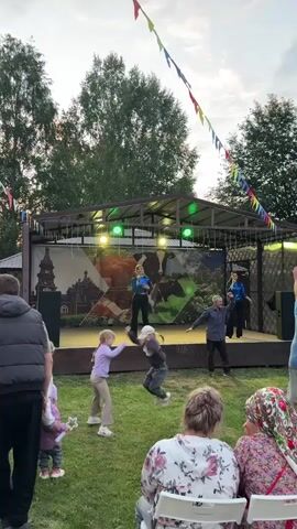 A group of people are gathered outdoors for an event. The focal point is a stage with performers, a singer, and dancers, with colorful lights and decorations. Children are dancing in front of the stage, interacting with each other. Adults are seated or standing, observing the performance. The environment appears to be a park or rural area with trees and open space.