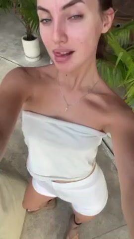A woman in a white tube top and white shorts poses for a selfie outside. She has light skin, blue eyes, and wears a silver necklace. She poses with pursed lips then winks. In the background is a stone surface, potted plants, and a cushion. Upbeat music plays.