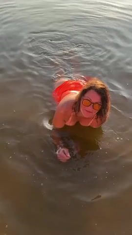 A woman with curly, auburn hair and sunglasses is in the water, wearing a coral-colored swimsuit. She is lying on her stomach and moving her legs, creating ripples in the water. The water is murky and reflects the sunlight. The scene is shot from a high angle, looking down at the woman.