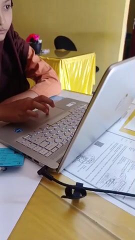 A young person with brown skin, wearing a dark brown hijab and an orange long-sleeved shirt, is seated at a yellow table, using a silver laptop. A blank white sheet of paper and a blue stapler are to the left of the laptop. To the right of the laptop are several sheets of paper containing printed text. A black power cord is plugged into the side of the laptop. The table is covered with a shiny yellow tablecloth. The background contains other yellow tables draped with similar tablecloths, and chairs. A plant in a pot is on one of the tables. The wall is painted yellow. The person appears to be focused on the laptop screen while typing.