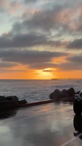 The video shows a coastal scene at sunset. In the foreground, a white Jeep Wrangler is parked on a wet, paved surface. The Jeep is partially obscured by the camera's perspective, with its front end and right side visible. Its top is down, revealing the interior. To the right of the Jeep, a black BMW M6 is parked, facing the ocean. Its headlights are on. The wet pavement reflects the lights of the BMW and the sunset. In the middle ground, the ocean waves crash against a rocky shoreline. In the background, the sun is setting over the horizon, casting a warm orange glow across the sky. Clouds are scattered across the sky, with some of them tinged with orange and pink from the sunset. A distant pier extends out into the ocean. The overall mood is serene and picturesque.