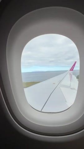 The view through an airplane window shows a wing and the horizon with an island. The wing is white with a purple detail on the end. The sky is cloudy and the water is choppy. The island is a light brown color with a slightly mountainous surface.