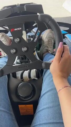 The video shows a first-person perspective of someone sitting in a go-kart at a racetrack. The person is wearing jeans and white sneakers. The steering wheel is black with carbon fiber accents. You can see the Sodi and Tekneex logos on the steering wheel. The racetrack is asphalt and has white directional arrows painted on it. There is another go-kart driving on the track. The background shows a building and a mountain range under a partly cloudy sky.