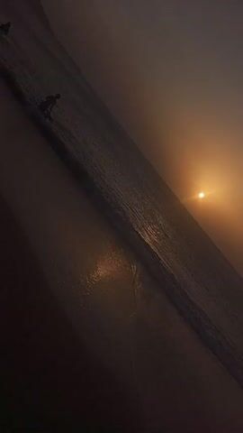 The video shows a beach at sunset. A lone figure is walking along the shoreline where the waves are breaking. The sun is low in the sky and casts a golden glow across the water and sand. The scene appears hazy and indistinct, with muted colors.