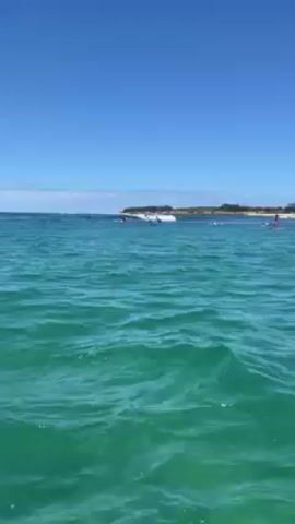 The video shows a sunny, clear day at the beach. The water is turquoise with gentle waves.  In the distance, a landmass with trees and a sandy beach is visible. Several people are in the water, some surfing and others paddleboarding.  The camera appears to be on a boat or some other vessel, bobbing slightly with the water.