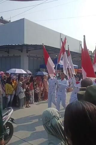 The video shows a parade with several individuals carrying flags that are red and white. They are marching in formation on a street lined with spectators. The individuals are wearing white uniforms and dark caps. Some spectators are holding umbrellas. The weather appears to be sunny. The parade is moving from left to right in the frame.