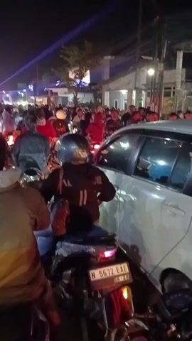 The video shows a crowded street at night, possibly during an event. Many people are on motorcycles, some are walking, and there is a white car in the foreground. Light beams are visible in the background, suggesting stage lighting or a projection. The motorcycles are stopped, indicating traffic or a gathering. One person in the foreground is wearing a helmet and a jacket. Another person in the foreground is wearing a brown jacket. The license plate on a motorcycle in the foreground reads "N 5672 EAE." The atmosphere appears lively and busy.