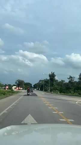 The video shows a view from a vehicle traveling on a road. The road has multiple lanes and is flanked by vegetation and trees. There are a few cars and a motorcycle on the road. The sky is cloudy. The overall environment appears to be rural or suburban.