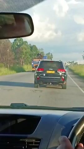 A driver is operating a car on a paved road. Ahead, a dark Jeep Grand Cherokee with license plate "ADE 4272" is driving. Further ahead, a large blue truck carrying heavy equipment is also driving. The sky is bright, suggesting daylight. Green trees line the roadside. The interior of the filming vehicle is visible, including the dashboard and the driver's hand on the steering wheel.