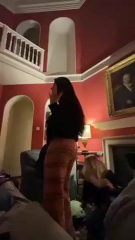 The video shows two people in an ornate room with high ceilings. One person, with long dark hair, is wearing a black top and orange striped pants, and they are dancing with a wine glass in their hand. The other person is bent over. The room is painted a deep red and features a fireplace, a painting, and a balcony.