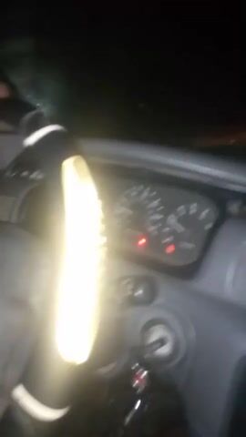 A person's hands grip a steering wheel in a vehicle at night. The steering wheel is covered with a reflective material, creating a bright, glowing effect. The dashboard instruments are visible, with lights illuminated. The overall scene is dark, indicating nighttime driving.