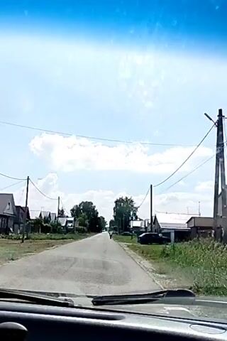 The video shows a view from inside a vehicle driving down a paved road through a village. The sun is shining and there are clouds in the sky. Houses are visible on both sides of the road. There are power lines running overhead. In the distance, a person is walking down the road.