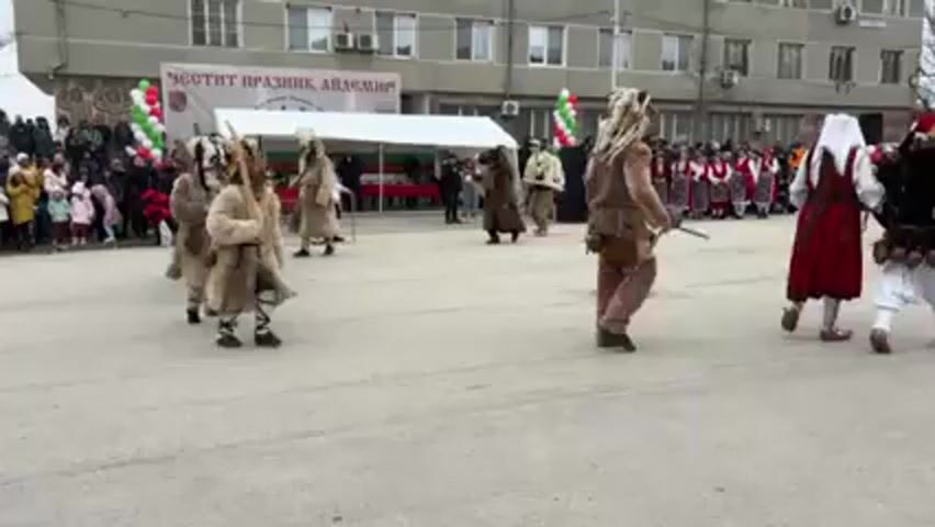 A procession of individuals dressed in traditional Bulgarian Koukeri costumes walks across a paved plaza. The costumes consist of thick, animal-fur coats, decorative belts, and elaborate horned masks, some with feathers. They are walking in a coordinated manner, and some carry wooden sticks. A crowd of spectators lines the left side of the plaza, many are children bundled in winter clothing. In the background is a building with a sign in Bulgarian and a white tent decorated with Bulgarian flags. The atmosphere suggests a festive, cultural event.