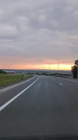 This is a first-person perspective video of a person driving on a highway during sunset. The road ahead is paved and has white lane markings. There are streetlights along the side of the road. In the distance, there are some vehicles, including trucks. The sky is cloudy with a gradient of colors from gray to orange and yellow, indicating the setting sun. The landscape on either side of the highway appears to be open land with some vegetation and a body of water in the distance. The video captures the motion of driving as evidenced by the blurred road and surroundings.