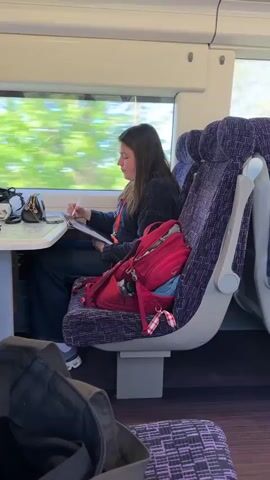 The image captures a woman seated in a modern train carriage, likely traveling during the daytime given the bright light streaming through the large window. The setting appears to be a train in Sevenoaks, United Kingdom.

The woman, with long, dark hair, is dressed in a dark sweater and dark trousers, and is wearing light-colored shoes. She is sitting comfortably at a table, facing slightly towards the window, and appears engrossed in an activity. She is holding a stylus-like object, possibly an Apple Pencil, and is writing or drawing on what looks like a tablet or a notebook. A red lanyard is visible around her neck, and she wears a watch on her left wrist.

On the table in front of her, there's a small black purse, a pair of sunglasses, and a white earbud case. Next to her on the adjacent seat rests a prominent red backpack, with the brand "Osprey" clearly visible on it. A blue reusable bag is tucked into the backpack, and a red and white checkered ribbon or keychain dangles from it. Part of a dark bag is visible in the foreground, suggesting other luggage.

The train seats are upholstered in a purple patterned fabric, and the interior walls are white, giving a clean and bright impression. Through the window, the outside scenery is blurred, indicating that the train is in motion. The overall atmosphere suggests a quiet and focused moment during a journey.