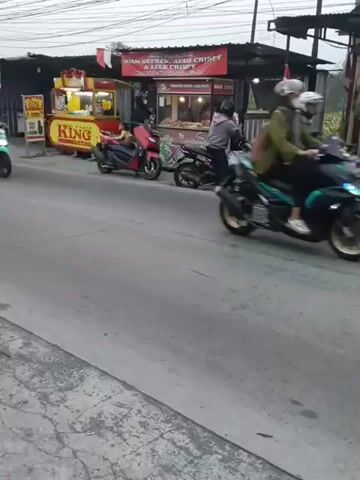 This is a video of a street scene with several motorcycles and food stalls. In the foreground, a black motorcycle with two passengers is traveling towards the viewer. The passenger in the back is wearing a light blue shirt and blue jeans, and the passenger in the front is wearing a brown jacket and a pink head covering. To the left, a teal-colored motorcycle with one rider is moving away from the camera. Further back, several other motorcycles are parked or moving. Behind the motorcycles are food stalls. One stall has a red sign that reads "AYAM GEPREK, AYAM CRISPY & LEK CRISPY". Another stall, to the left of that, has a yellow and red sign with the word "KING" visible. The stalls appear to be open, with food being prepared or sold. The environment appears to be an urban or semi-urban setting with overhead wires visible. The lighting suggests it is either early morning or late afternoon.