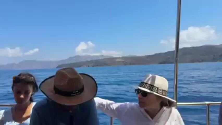 Three people are seated on a boat on a clear day. The woman on the left wears sunglasses and a light-colored top. The person in the middle wears a straw hat with a black band and a dark shirt. The woman on the right wears a white hat with a Greek key design, sunglasses, and a white shirt. She points towards the land in the background, which appears to be a mountainous island. The sea is blue with small waves, and the sky is mostly clear with some white clouds. The boat has a metal railing.