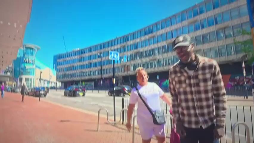 A man wearing glasses, a baseball cap, and a plaid shirt stands on a sidewalk in front of a large, modern building with many windows. To his left, a woman in a white t-shirt and shorts, with her hair in a bun, is looking down at a pink bag. Behind them, cars drive down a street, and further in the background, a blue sign with a bicycle symbol is visible on the building. The sky is clear and blue.