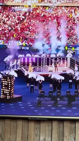 A concert stage is crowded with performers, many holding white umbrellas. The background is filled with a dense crowd. The stage is lit with colorful lights, and smoke effects are being used. A close-up shot then focuses on a singer at a microphone. The lighting changes from blue to green as the singer performs.