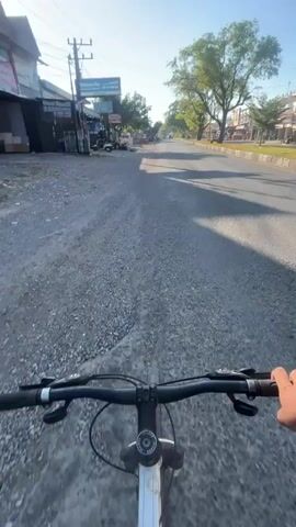 The video shows a first-person perspective of someone riding a bicycle down a paved road. The sun is shining brightly, casting long shadows. On the left side of the road, there are buildings, some with awnings, and parked motorcycles. There are also utility poles and wires. On the right side of the road, there is a curb, a sidewalk, more buildings, and trees. The road itself is mostly empty, with the exception of some distant vehicles. The rider's hands are visible on the handlebars of the bicycle. The overall environment appears to be a town or city street during daylight hours, possibly in the morning or late afternoon due to the low angle of the sun.
