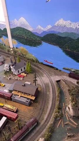 The video pans across a meticulously detailed model train set. The landscape features a winding blue river, bordered by lush green hills and snow-capped mountains under a bright blue sky. A small red canoe with two figures is visible on the river. The foreground depicts a quaint town with miniature buildings, including a church with a steeple, houses, shops, and a gas station. Several model trains are positioned on the tracks, some appearing to be in motion. The scene also includes various miniature vehicles, trees, streetlights, and tiny human figures engaged in everyday activities, such as standing around the church and by the train station. The overall impression is a vibrant, miniature representation of a town nestled in a scenic natural environment.