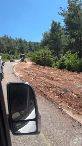 The media file shows a view from the passenger side of a vehicle, looking towards a road under construction. The scene is set in a rural, mountainous area with lush green trees lining the road. The weather appears to be sunny and clear, as indicated by the bright blue sky.

The primary object of interest is a grader, a large piece of construction equipment, actively working on the road. The grader is moving dirt and debris, suggesting road repair or widening work is in progress. A pile of reddish-brown dirt is visible to the right of the road, likely a result of the grading operation. Other vehicles, including cars and possibly another construction vehicle, are visible in the distance on the road, indicating ongoing traffic despite the construction.

The foreground of the image is dominated by the side mirror of the vehicle the footage is being filmed from, reflecting a portion of the vehicle's interior and exterior. The road itself is asphalt, with yellow lines marking the lanes. The surrounding landscape is natural and undeveloped, with dense foliage and trees. The overall impression is one of a typical day in a somewhat remote area where infrastructure development is taking place. No people are clearly visible, but their presence is implied by the construction activity. The time of day is likely midday due to the bright sunlight casting strong shadows. The location cues, such as the vegetation and the type of terrain, along with the provided location context of Osmaniye, Turkey, suggest a Mediterranean or similar climate. There is some visible text on the side of the grader, but it is not clear enough to be read.