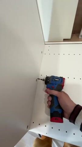 A person's right hand is shown holding a blue and red/black Vinssok brand power drill. The drill, which has a "BL" marking on its battery pack, is being used to drive screws into white cabinet or furniture panels. The drill's integrated light illuminates the area where the screw is being inserted. The person is fastening screws into the interior corner of what appears to be a cabinet frame, with several other screws already present along the edges of the white panels. The background shows additional white furniture components.