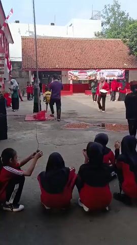 A group of people, primarily children and adults, are gathered in an outdoor courtyard. The courtyard appears to be part of a school or community center, with a building featuring red and white accents and a terracotta tiled roof in the background. A flag pole stands in the center of the courtyard, and a banner with text and images is displayed on the building. Many of the children are wearing red and black uniforms, including headscarves for some. An adult in a purple shirt is near the flag pole, possibly involved in a ceremony. Another adult in a red shirt and headscarf is standing and holding papers. Several children are seated on the ground in the foreground, facing the activity. Other children are standing in lines or groups, some appearing to be participating in an activity or watching. The ground is paved, and there are puddles of water in some areas. Trees are visible in the background. The overall atmosphere suggests an organized event, possibly a flag-raising ceremony or a school celebration.