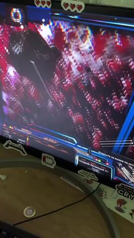 A child in a red and blue hooded robe is sitting at a wooden desk with a black keyboard, looking at a computer monitor covered in stickers. The monitor displays a video game with a red and black space scene. There are some papers and a blue pen on the desk.