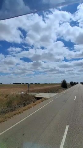 The video shows a view from a vehicle traveling on a paved road. The road has a white dashed line marking the center and a solid white line on the right edge. On the left side of the road, there is a dry, grassy field. On the right side, there are more fields and some trees. The sky is blue with scattered white clouds. The sun appears to be shining, creating some glare on the windshield. There are no people visible in the video. The overall environment suggests a rural setting on a clear day.