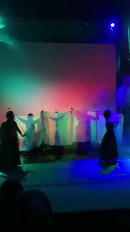 The video shows a theatrical performance. Several dancers in traditional costumes perform on a stage with a large screen in the background displaying shifting colored lights and mist. Stage lights of various colors, including red, green, and blue, illuminate the stage and dancers. An audience is visible in the foreground, with focus on the performers and stage setup. The lighting effects add a dramatic atmosphere to the performance.