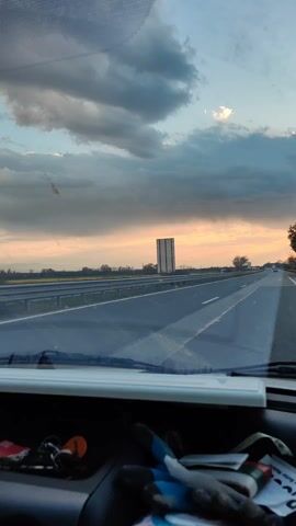 The video, shot from inside a vehicle, shows a drive on a highway under a cloudy sky at sunset.  The windshield shows a dashboard with keys and other items. On the driver's side, the side mirror reflects the passing landscape of fields and trees.  Other vehicles can be seen further up the road.