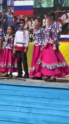 A group of children, approximately five to seven, are performing on a blue wooden stage. The girls wear bright, colorful traditional dresses with pink skirts and patterned tops. The boys are in white shirts with red trim and black pants. Microphones are set up in front of them. They are singing or reciting something. Behind the stage is a backdrop with images of people in traditional clothing and a Russian flag. The setting appears to be an outdoor event on a sunny day.
