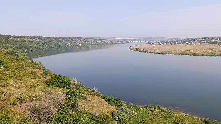 A drone shot pans across a wide, calm river with a slight haze in the sky. The riverbanks are lined with green vegetation, including trees and bushes. On the left side of the river, a steep, vegetated hill slopes down to the water. On the right side, a flat, dry, golden field stretches towards a small village with scattered buildings and a bridge visible in the distance. The water reflects the sky and the surrounding landscape.