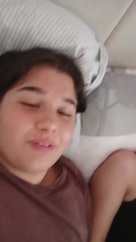 The video shows a young girl in a bedroom. The first shot is a close up of a girl’s face. She is wearing a brown shirt. The next shot is a wider view of the bedroom. Another girl is lying on the bed, looking at a cell phone. She is wearing a white shirt and dark pants. A small white dog is also visible near the foot of the bed. The last shot is another close up of the first girl’s face.