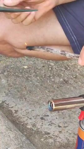 A person's hands hold a piece of glass and a stick that is being heated by a torch. The stick is burning and producing smoke. The background is an outdoor setting with a concrete surface. The person is wearing blue shorts.
